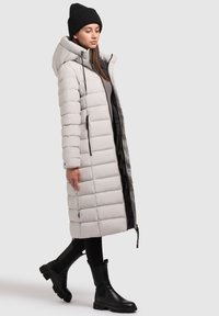 Lange, lichtgrijze pufferjas met horizontale stiksel, een capuchon, een rits aan de voorkant en zijzakken, gedragen over zwarte leggings en enkelboots.