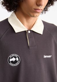 Mørk grå polo-shirt med en cremefarvet krave, der har et cirkulært broderet logo på brystet og en knaplukning. Blødt, struktureret stof.