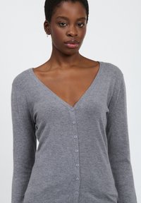 Grauer Cardigan mit langen Ärmeln, V-Ausschnitt und Knopfverschluss auf der Vorderseite. Weicher Stoff und dezentes Strickmuster.