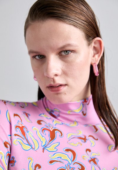 Roze blouse met hoge hals en een patroon, met abstracte ontwerpen in rood, geel en blauw. De accessoires omvatten roze geometrische oorbellen.