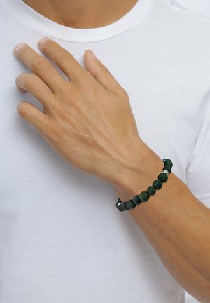 Männliche Hand mit dunklem Perlenarmband auf der Brust ruhend, trägt schlichtes weißes T-Shirt mit Rundhalsausschnitt.