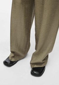 Pantalones anchos de color verde oliva en una tela texturizada, combinados con zapatos negros de punta cuadrada que presentan acentos brillantes y mate.