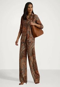 Γυναίκα που φοράει ένα καφέ jumpsuit με σχέδιο paisley και κρατάει μια μεγάλη καφέ δερμάτινη τσάντα στον ώμο.