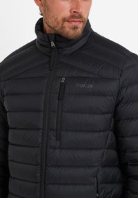 Black puffer jacket with a high collar, quilted texture, and zippered pockets. Features subtle branding on the chest.