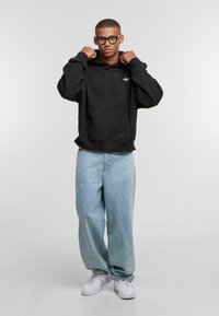 Black hooded sweatshirt with front pocket, loose fit, paired with light blue baggy jeans and white sneakers, on a plain background.