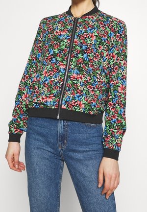 Femme portant une veste bomber zippée à fleurs colorées avec des poignets et un col noirs, associée à un jean bleu taille haute.