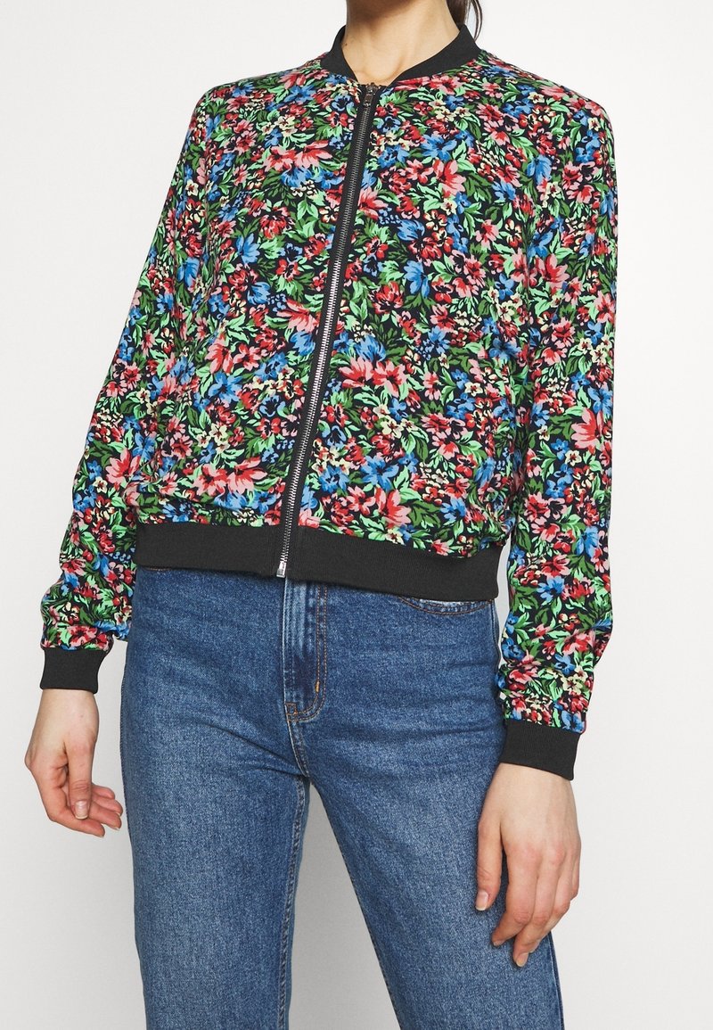 Femme portant une veste bomber zippée à fleurs colorées avec des poignets et un col noirs, associée à un jean bleu taille haute.