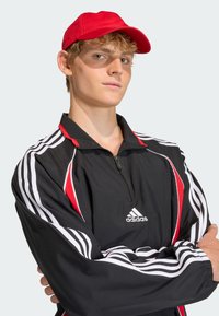 Čierna športová bunda na zips s červenými akcentmi a bielymi pruhmi, doplnená červenou šiltovkou. Vyznačuje sa hladkou látkou a štruktúrovaným strihom.