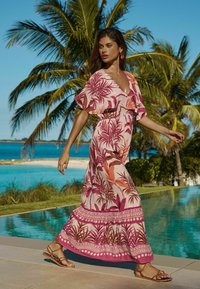 Rochie maxi cu imprimeu de frunze de palmier roz și portocalie, mâneci scurte, decolteu adânc în V, talie strânsă și tiv cu model; purtată cu sandale maro.