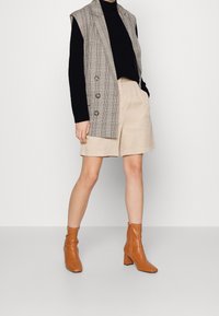 Grauer karierter ärmelloser Blazer über einem schwarzen Rollkragenpullover, kombiniert mit beigen Shorts und braunen Stiefeletten mit einem blockigen Absatz.