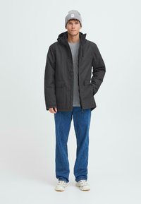 Svart parkasjakke med frontlommer, justerbar hette og glidelås, iført en grå genser og blå jeans. Modellen står oppreist.