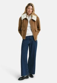 Braune Wolljacke mit weißem Schaffellkragen, ausgestattet mit zwei Fronttaschen, getragen über einem weißen Strickpullover und blauen weit geschnittener Jeans.