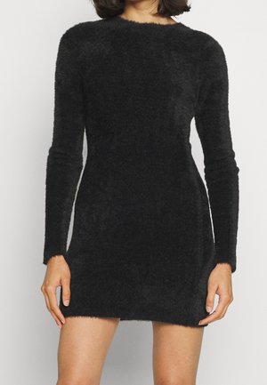 Frau trägt ein langärmliges, schwarzes, flauschiges Strick-Minikleid und steht vor einem schlichten hellgrauen Hintergrund.