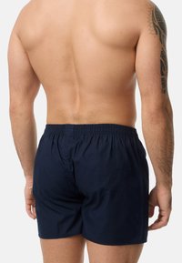 Námořnické modré plavkové šortky s elastickým pasem, hladkou texturou, délkou ke kolenům a minimálními detaily šití, které nosí mužský model s odkrytým zády.