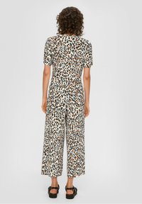 Osoba z kręconymi włosami stoi tyłem, ubrana w bluzkę z krótkim rękawem w panterkę i szerokie spodnie, a na stopach ma czarne sandały, na jednolitym tle.