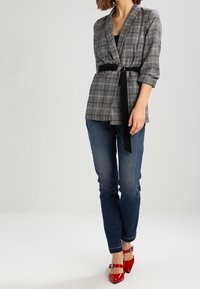 Mujer vestida con un blazer de cuadros gris con cinturón negro, jeans azules y tacones rojos de punta, de pie contra un fondo claro.