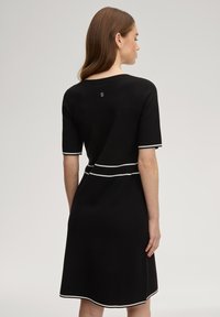 Μαύρο πλεκτό φόρεμα με κοντά μανίκια, διακοσμημένο με λευκές λεπτομέρειες στα μανσετάκια και στη μέση, με στρογγυλή λαιμόκοψη και μικρό λογότυπο στο πίσω μέρος.