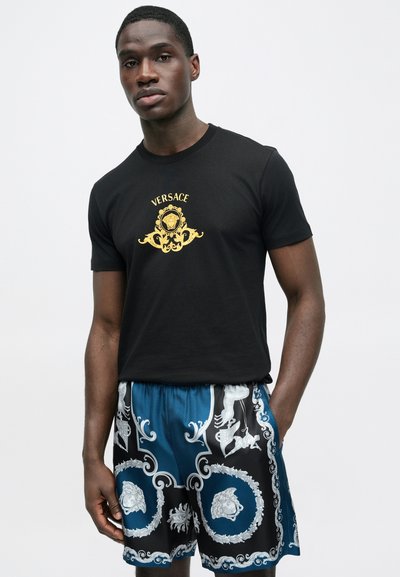 Tricou negru din bumbac cu logo-ul auriu „VERSACE”, asortat cu pantaloni scurți albaștri și negri, având modele albe complexe și talie cu șnur.