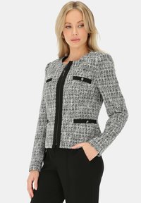 Szara tweedowa marynarka z czarnymi akcentami, zapinana na zamek, z dwoma kieszeniami na piersi i teksturowaną tkaniną. Dopasowany krój i długie rękawy.