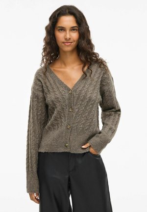 Bruine kabelbrei cardigan met een V-hals, voorzien van gouden knopen en lange mouwen, gecombineerd met zwarte pants voor contrast.