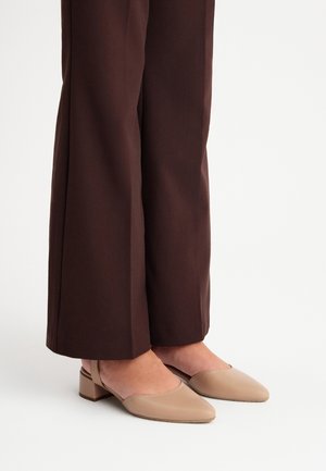 Osoba nosząca brązowe spodnie o szerokich nogawkach i beżowe sandały typu slingback na niskim klocku na białym tle.