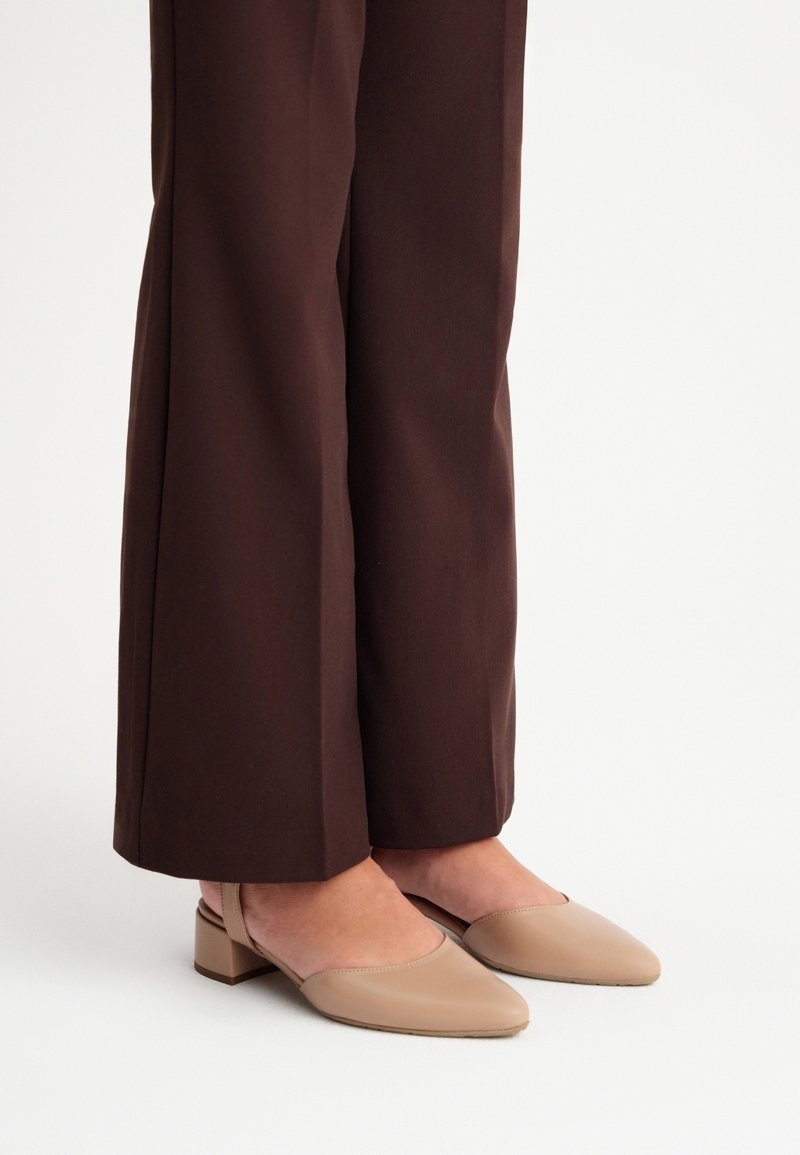 Personne portant un pantalon marron à jambes larges et des chaussures slingback beige à petit talon bloc sur fond blanc.