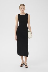 Black sleeveless dress with a fitted silhouette, knee-length, smooth texture. Model holds a cream bag and wears black flip-flops.