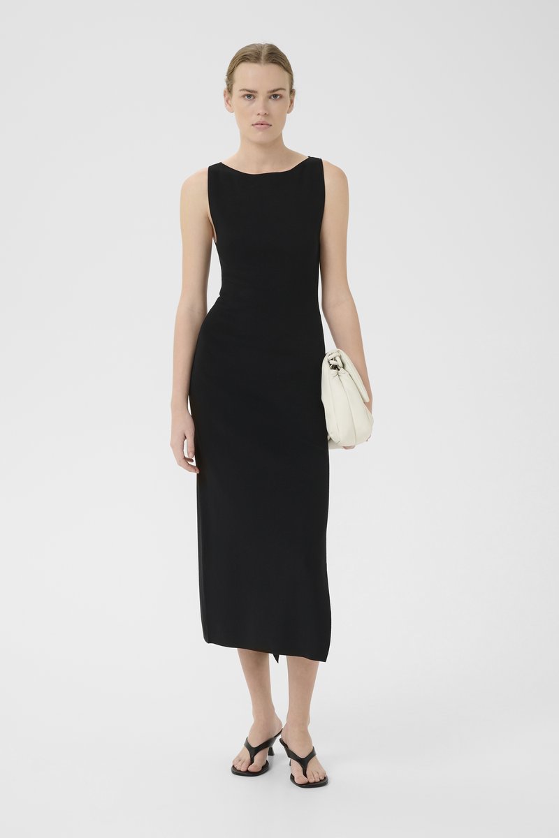 Black sleeveless dress with a fitted silhouette, knee-length, smooth texture. Model holds a cream bag and wears black flip-flops.