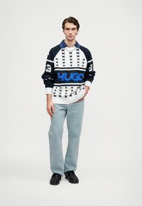 Pulover ima črno-belo vzorje z modrim logotipom "HUGO", v kombinaciji s svetlo modrimi hlačami in črnimi čevlji. Teksturovan pleteninski material.