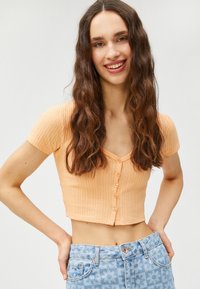 Kurzärmliges, geripptes Crop-Top in hellorange mit V-Ausschnitt und fünf Knopfverschluss vorne; kombiniert mit hellblauen Jeans mit floralem Muster.
