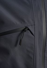 Mörk, texturerad tyg med vertikala ribbor. Har en ficka med dragkedja med en kontrasterande svart dragkedja och detaljer längs sömmarna.