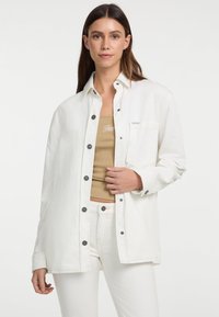 Witte overhemdbloes gemaakt van denim met zwarte knopen, voorzien van een borstzak en een relaxte pasvorm, gedragen over een beige tanktop.