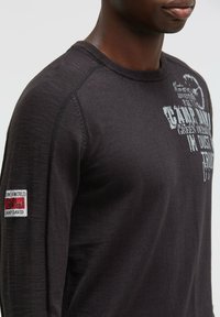 Dunkelbluse mit langen Ärmeln aus strukturiertem Stoff; enthält weißen grafischen Text und ein aufgenähtes Patch mit roten Akzenten am linken Ärmelt.