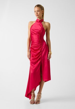 Femme debout portant une robe en satin rouge froncée sans manches, avec un col montant, un ourlet asymétrique et des talons dorés à brides ainsi que des bracelets.