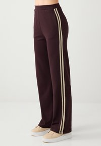 Pantalon large bordeaux avec des bandes latérales crème. Confectionné dans un tissu doux, doté d'une taille élastique et d'une coupe confortable. À associer avec des baskets beige.