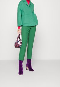 Veste en mélange de laine verte avec un col et deux poches avant, assortie à un pantalon ajusté vert. Les chaussures sont des bottines en velours violet.