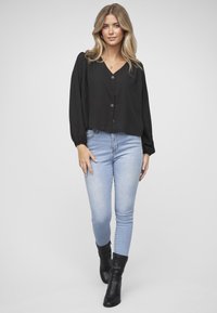 Blusa negra abotonada con escote en V y mangas largas abullonadas. Combinada con vaqueros pitillo azul claro y botines negros. Fondo neutro.