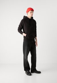 Melnā kapuce ar priekšējo kabatu un regulējamām auklām, apvienota ar tumšiem džinsiem un melniem apaviem. Uz galvas sarkans adīts beanie.