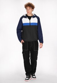 Jachetă ușoară cu fermoar, cu panouri bleumarin, negru și alb, o dunga albastră, glugă ajustată și manșete elastice, purtată cu pantaloni cargo negri.