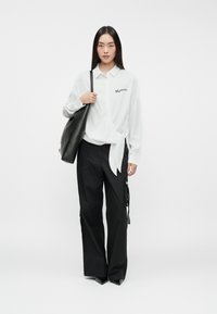 Camisa blanca de manga larga con un lazo en la cintura, combinada con pantalones negros holgados con cremalleras y un parche de logo, y una bolsa tote negra.
