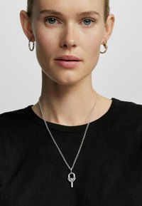 Silberne Kettenhalskette mit einem einzigartigen Anhänger in Schlaufen- und Stabdesign, getragen über einem schwarzen T-Shirt mit Rundhalsausschnitt.