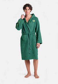 Groene badjas met een gestructureerde stof, lange mouwen, twee zakken aan de voorkant en een ceintuur. Heeft een logo op de borst en een geborduurd detail op de mouw.