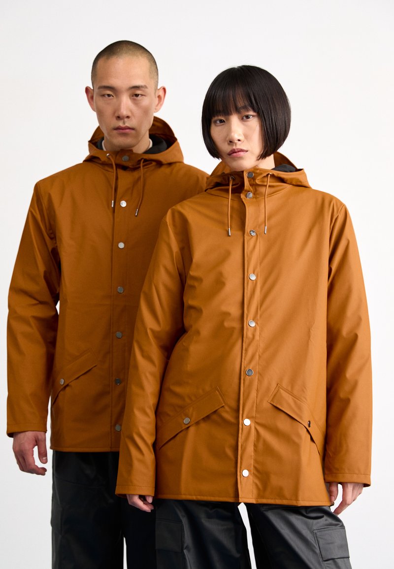 Jachete impermeabile maro cu glugă, dotate cu închideri cu capse și buzunare laterale. Textură netedă, design unisex, purtate de doi modele.