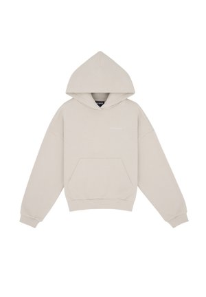 Personne portant un sweat à capuche beige, un pantalon large, des chaussures beiges, une casquette bleu marine, tenant un blazer beige assorti sur un fond gris clair.