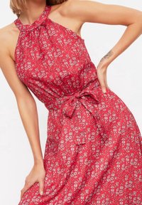 Kvinna i ärmlös röd klänning med vitt blommönster och halterneck, knuten med bälte i midjan, poserande med en hand på höften.