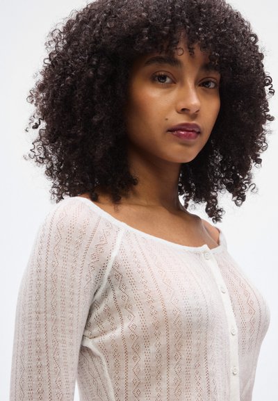Λευκό πλεκτό ζακέτα με υφασμένο σχέδιο, με στρογγυλή λαιμόκοψη και κούμπωμα με κουμπιά. Μαλακό υλικό και εφαρμοστή σχεδίαση, μακριά μανίκια.
