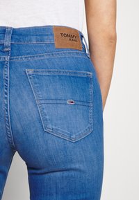 Jeans de mezclilla azul con una etiqueta de piel marrón de TOMMY JEANS en la parte posterior. Presenta bolsillos traseros con detalles de costura. Textura suave.