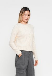 Giovane donna con lunghi capelli castani e biondi, che indossa un maglione lavorato a trecce color crema e pantaloni cargo grigi, con le mani nelle tasche.