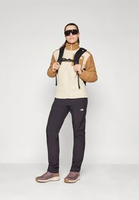 Kreemikas fliisjakk pruunide detailidega, mustad matkafirmad püksid ja pruunid matkasaapad. Kandjal on päikeseprillid ja must seljakott.