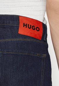 Mörkblå denimjeans med en röd läderetikett märkt "HUGO" vid midjan. Har guldtrådar och en silvermetallaccent.